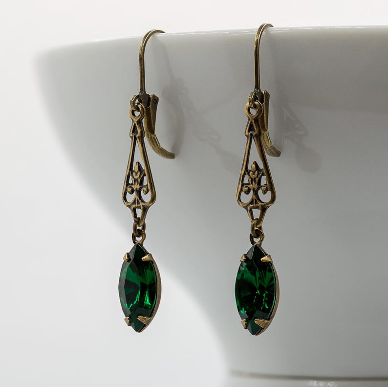 Pu&ograve; includere: Un paio di orecchini color oro antico con un design a filigrana. Ogni orecchino presenta una pietra preziosa verde smeraldo intenso a taglio marquise. Gli orecchini hanno una chiusura a gancio e un'estetica vintage.