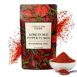 May include: A package of Korean red pepper flakes, labeled "Collected Foods." The package has a red and green design. A small white bowl and spoon contain the red pepper flakes. The net weight is 2 oz (56g).