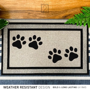 May include: A beige doormat with a black border and three black paw prints. The text "WEATHER RESISTANT DESIGN" and "BOLD & LONG LASTING UV INKS" is printed on the bottom of the mat.