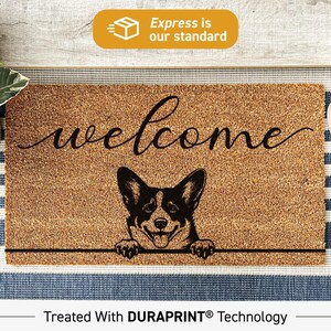 May include: A rectangular tan coir doormat featuring the word welcome in black cursive script above a black and white illustration of a Corgi dog with its front paws resting on a horizontal line. The mat is treated with DURAPRINT technology.