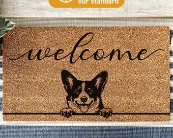 May include: A rectangular tan coir doormat featuring the word welcome in black cursive script above a black and white illustration of a Corgi dog with its front paws resting on a horizontal line. The mat is treated with DURAPRINT technology.