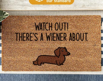 May include: A rectangular brown coir doormat featuring the black text WATCH OUT! THERE'S A WIENER ABOUT. and a graphic illustration of a brown dachshund dog. The mat is treated with DURAPRINT technology for durability and clear design visibility.