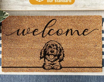 Könnte beinhalten: Eine rechteckige, hellbraune Kokosfasermatte mit dem Wort welcome in schwarzer Schreibschrift über einer schwarzen Strichzeichnung eines flauschigen Hundes. Der Hund ist unten mittig platziert und stützt seine Vorderpfoten auf eine schwarze Linie.
