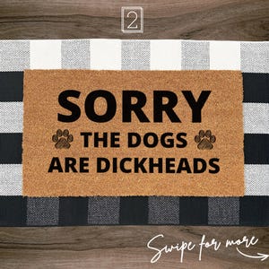 May include: A rectangular tan coir doormat featuring the black text SORRY THE DOGS ARE DICKHEADS with a paw print icon on each side of the middle line. The mat is layered over a black and white buffalo check rug with a square border pattern.