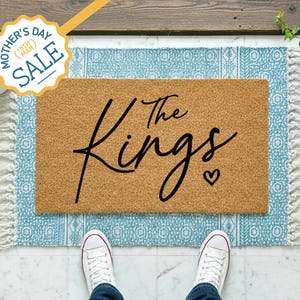 May include: A rectangular doormat with the words "The Kings" in black script and a small heart. The mat is on a blue patterned rug with white fringe. The image includes a Mother's Day Sale badge.