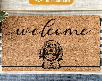 May include: A brown coir doormat with the word "welcome" in black cursive script. A black silhouette of a dog's head is peeking over the top of the mat. The text "Treated With DURAPRINT® Technology" is printed at the bottom of the mat.