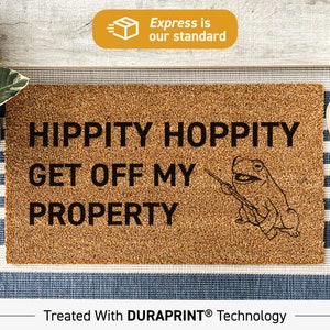 Op de afbeelding: Een bruine kokosvezel deurmat met de tekst "Hippity Hoppity Get Off My Property" en een cartoon kikker die een stok vasthoudt. De deurmat is behandeld met Duraprint-technologie.