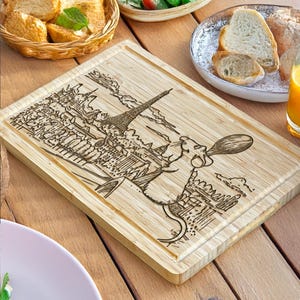 Puede incluir: Tabla de cortar rectangular de bamb&uacute; con un dise&ntilde;o grabado con l&aacute;ser de la Torre Eiffel y un chef de dibujos animados sosteniendo una cuchara. La tabla es de color marr&oacute;n claro y tiene un borde biselado. El dise&ntilde;o es marr&oacute;n oscuro.