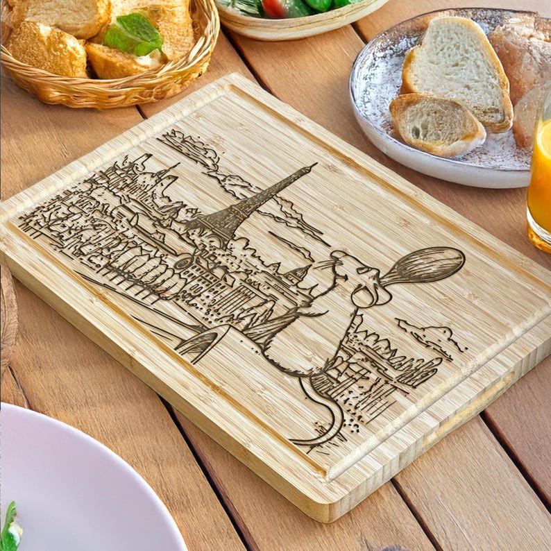 Puede incluir: Tabla de cortar rectangular de bamb&uacute; con un dise&ntilde;o grabado con l&aacute;ser de la Torre Eiffel y un chef de dibujos animados sosteniendo una cuchara. La tabla es de color marr&oacute;n claro y tiene un borde biselado. El dise&ntilde;o es marr&oacute;n oscuro.