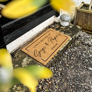 Puede incluir: Una alfombra de coco marr&oacute;n con el texto "Life's better at Cygi & Papa's House" en letras negras.