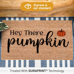 Puede incluir: Una alfombra de coco marrón con el texto "Hey There pumpkin" en fuente cursiva negra. Una pequeña calabaza naranja con un tallo verde está a la derecha del texto. La alfombra está tratada con tecnología DURAPRINT.