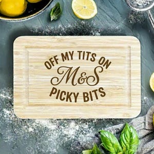 Può includere: Tagliere rettangolare in legno con la scritta "OFF MY TITS ON M&S PICKY BITS" in lettere marroni. La tavola è circondata da limoni, foglie di basilico e un setaccio.