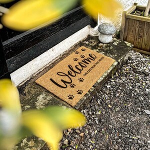 Op de afbeelding: Een bruine kokosmat met de tekst "Welcome" en "The cats run this house" in zwarte letters. De mat heeft zwarte poot afdrukken.