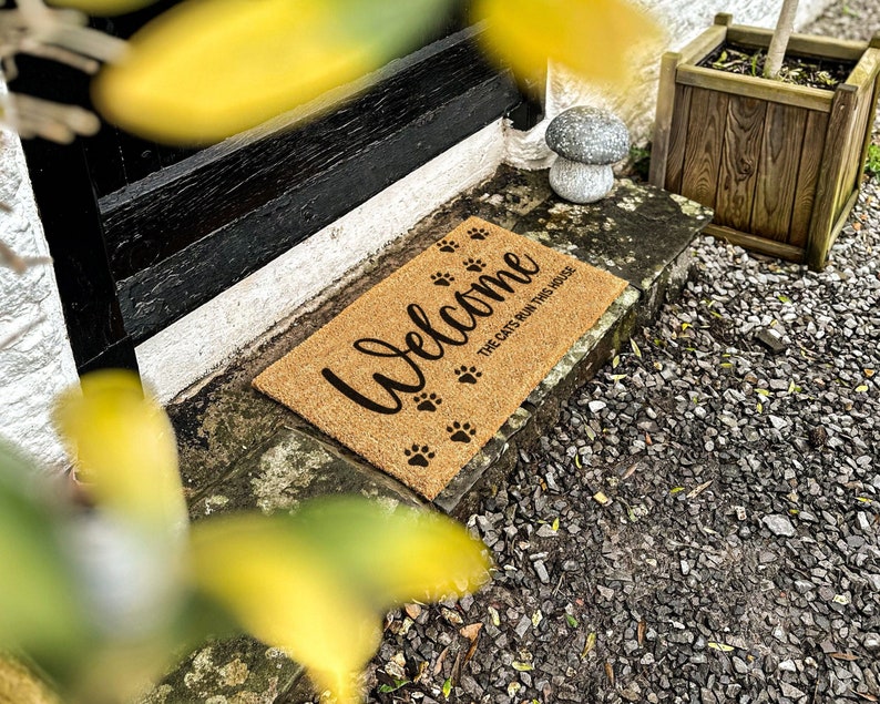 Op de afbeelding: Een bruine kokosmat met de tekst "Welcome" en "The cats run this house" in zwarte letters. De mat heeft zwarte poot afdrukken.