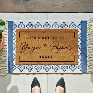 May include: A rectangular doormat with the text "LIFE'S BETTER AT Yaya & Papa's HOUSE" in black script. The mat is brown with a black border, and is surrounded by a blue and white patterned border with fringe.
