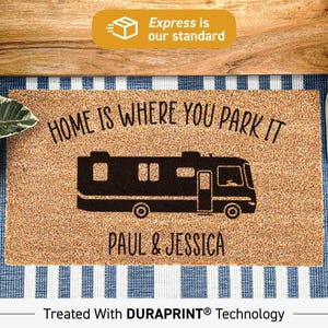 Puede incluir: Un felpudo rectangular de fibra de coco color canela con un gráfico negro de un vehículo recreativo. El texto HOME IS WHERE YOU PARK IT aparece sobre el vehículo y PAUL & JESSICA debajo. Tratado con la tecnología DURAPRINT.
