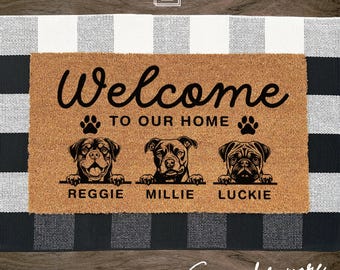 May include: A rectangular tan coir doormat featuring the black text Welcome TO OUR HOME. Below the text are three illustrated dog heads with paws resting on lines, labeled REGGIE, MILLIE, and LUCKIE, with two black paw prints above the dogs.