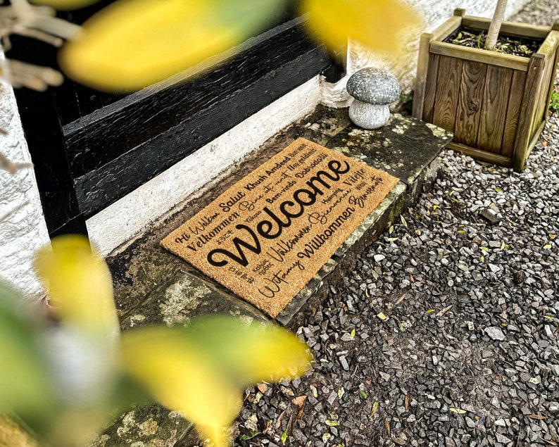 May include: A brown coir welcome mat with the word "Welcome" in large black lettering. The mat also features the word "Welcome" in multiple languages, including "Bem-vindo", "Hi Wilkin", "Salut", "Khush Amdeed", "Velkommen", "Be", "Witamy", "Willkommen", "B", "Vilejte".