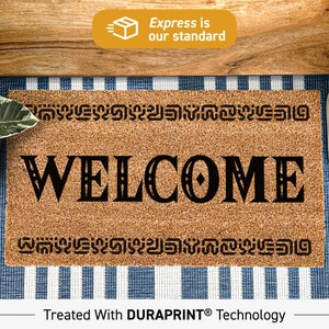 Peut inclure: Un paillasson en fibre de coco brun avec le mot "WELCOME" en lettres stylisées noires. Le paillasson a une bordure décorative et le texte "Treated With DURAPRINT® Technology".