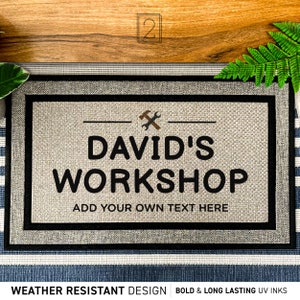 May include: A gray doormat with black trim and the text "DAVID'S WORKSHOP" in black. The text "ADD YOUR OWN TEXT HERE" is also in black. The doormat has a weather-resistant design and bold, long-lasting UV inks.