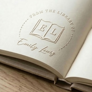 Op de afbeelding: Open boek met reliëftekst en een cirkelvormig ontwerp met de tekst "FROM THE LIBRARY OF" boven de naam "Emily Lewis". Het ontwerp bevat een open boek met de letters "E" en "L". Het boek heeft een beige kaft.