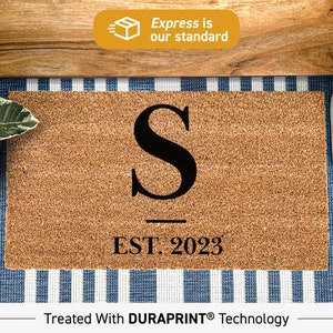May include: A rectangular doormat with a brown coir surface. A large black letter "S" is centered, with "EST. 2023" below. The text "Express is our standard" is in a yellow banner. The mat is on a blue and white striped surface.