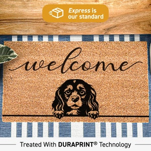 Op de afbeelding: Een bruine kokosmat met het woord "welcome" in zwarte cursieve letters. Een zwart silhouet van een Cavalier King Charles Spaniel-hond kijkt over de bovenkant van de mat. De mat ligt op een blauw-wit gestreept oppervlak. De tekst "Treated With DURAPRINT® Technology" staat onderaan de afbeelding.