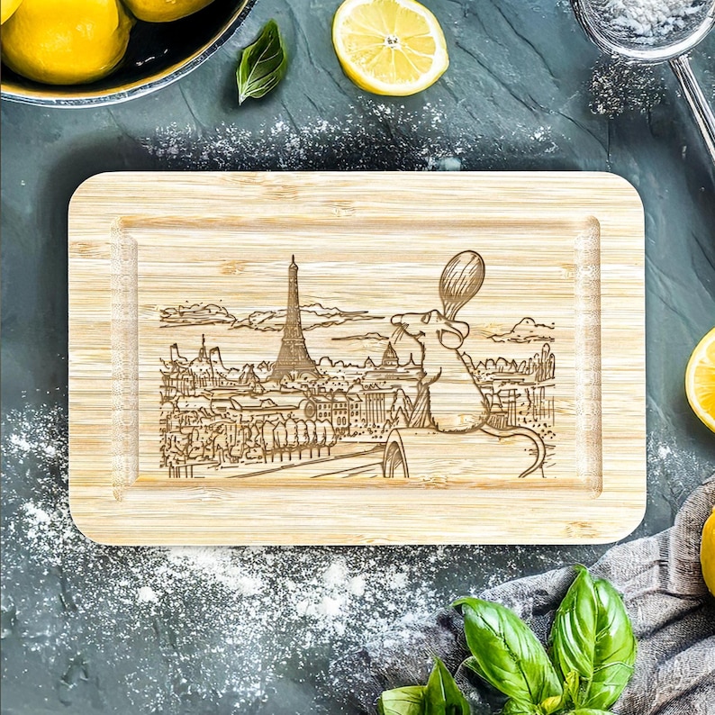 Puede incluir: Tabla de cortar rectangular de bamb&uacute; con un dise&ntilde;o grabado con l&aacute;ser de la Torre Eiffel y un rat&oacute;n de dibujos animados sosteniendo una cuchara. La tabla tiene una veta de madera clara y un borde empotrado. El dise&ntilde;o es de estilo lineal detallado.