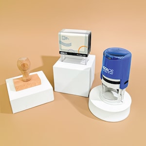 May include: Three different rubber stamps on white platforms. The first stamp is a wooden stamp with a rectangular base. The second stamp is a clear plastic stamp with a blue and white design and the text "2 Day Gifts Printer 40". The third stamp is a blue rubber stamp with the text "COLOP Printer 40".