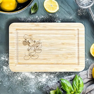 Könnte beinhalten: Ein Schneidebrett aus Holz mit einem Mickey-Mouse-Design. Das Brett ist mit einem Cartoon-Bild von Mickey Mouse als Koch graviert, der einen Topf und einen Löffel hält. Das Brett ist hellbraun und rechteckig.