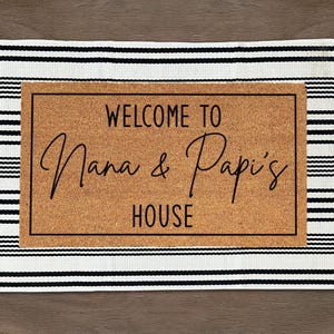 May include: Rectangular doormat with a brown center and a black and white striped border. The center features the text "WELCOME TO Nana & Papi's HOUSE" in a stylish script font.