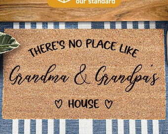 May include: A rectangular tan coir doormat featuring black text that reads There's No Place Like Grandma & Grandpa's House with a heart icon on each side of the word House. The mat is treated with Duraprint technology for a durable finish.