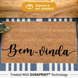 Puede incluir: Una alfombra de coco marrón con el texto "Bem-vinda" en letras cursivas negras. La alfombra está sobre una superficie a rayas azules y blancas. El texto "Treated With DURAPRINT® Technology" está en la parte inferior de la imagen.