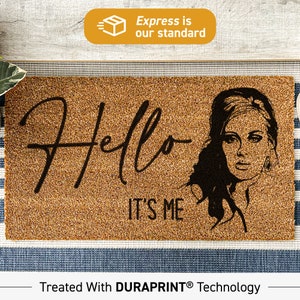 May include: A brown coir doormat with the text "Hello It's Me" in black cursive font. A black and white illustration of a woman with a bouffant hairstyle is on the right side of the doormat.