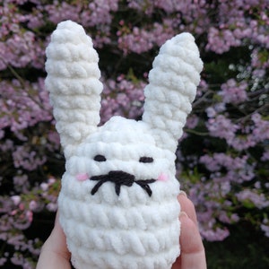 May include: A white, plush, crocheted bunny toy with long ears, black embroidered whiskers, and pink cheeks. The bunny is held in front of a background of pink blossoms.
