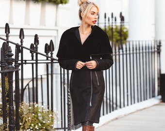 May include: A woman wearing a black long-sleeved coat with a fur collar and brown knee-high boots. She is walking down a sidewalk in front of a white building.