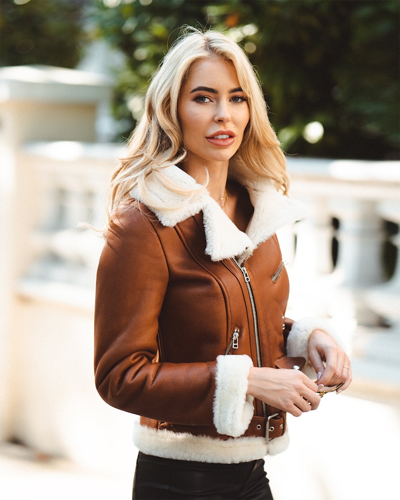 May include: A brown leather jacket with a white shearling collar and lining. The jacket has a zipper closure and a belt at the waist.