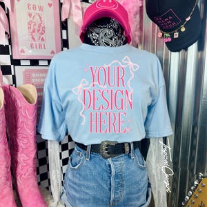 May include: A light blue t-shirt with the text "YOUR DESIGN HERE" in pink, paired with denim shorts and a black belt. The display includes a pink cap, a black trucker hat, and pink cowboy boots. The background features framed art and a checkered pattern.