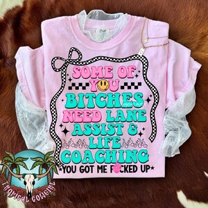 May include: A light pink t-shirt with the text "SOME OF YOU BITCHES NEED LANE ASSIST & LIFE COACHING YOU GOT ME FUCKED UP" in a retro font. The shirt has a lace collar and sleeves. A gold necklace with a star pendant is draped over the shirt.