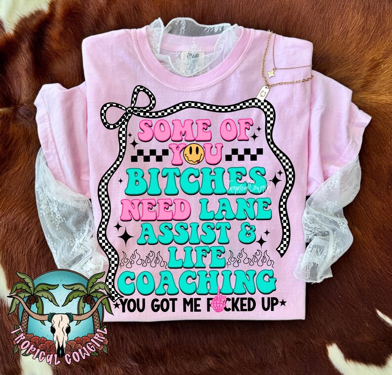 May include: A light pink t-shirt with the text "SOME OF YOU BITCHES NEED LANE ASSIST & LIFE COACHING YOU GOT ME FUCKED UP" in a retro font. The shirt has a lace collar and sleeves. A gold necklace with a star pendant is draped over the shirt.
