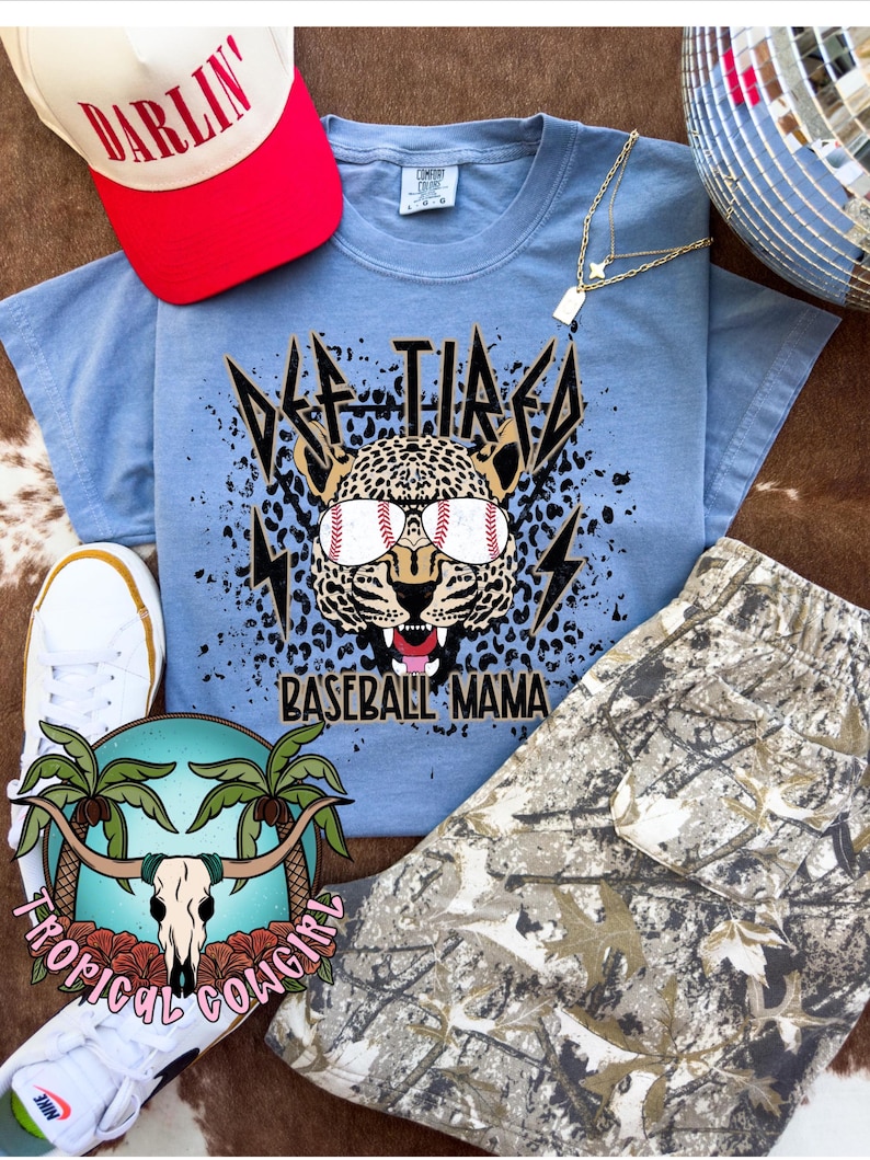 May include: A light blue t-shirt with a graphic of a leopard wearing sunglasses and baseballs. The text "Def Tired Baseball Mama" is printed below the graphic. The t-shirt is laying on a camouflage fabric.