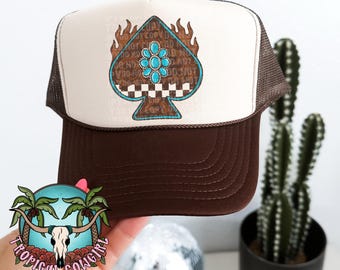 May include: Brown and white trucker hat with a brown brim and a white mesh back. The front of the hat features a brown and white spade design with a turquoise and white flower in the center. The spade design has the text "Do Not Copy Do Not Sell" printed on it.