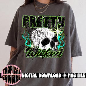 Può includere: Una t-shirt grigio antracite con una stampa grafica di un teschio bianco dalla texture usurata. Il testo PRETTY si trova sopra il teschio e Wicked sotto in lettere nere con contorni verde neon, circondato da fiamme color ottanio.