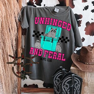 May include: A dark gray t-shirt with the text "Unhinged and Feral Moms Club" in pink and black lettering. A cartoon image of raccoons sitting on a blue cooler is featured on the shirt.