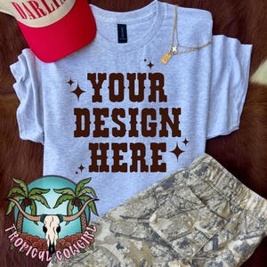 Könnte beinhalten: Ein hellgraues T-Shirt mit dem Text "YOUR DESIGN HERE" in braunen Buchstaben. Das Shirt liegt flach auf einer braunen Oberfläche mit einer weißen und roten Baseballkappe, einer goldenen Kettenkette und einer Tarnhose.
