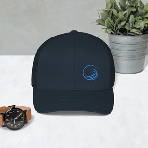 May include: A navy blue trucker hat with a blue wave logo embroidered on the front. The hat has a mesh back and a curved brim.