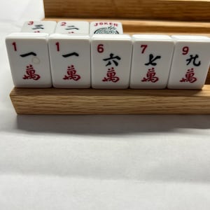 May include: A set of white mahjong tiles with red Chinese characters and numbers. The tiles are arranged in a row on a wooden rack. The tiles include the numbers 1, 1, 6, 7, and 9.