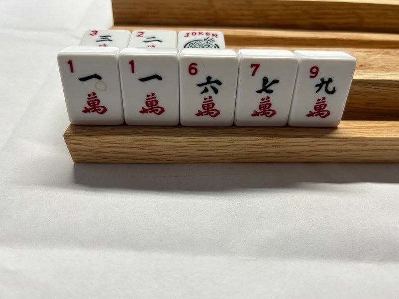 May include: A set of white mahjong tiles with red Chinese characters and numbers. The tiles are arranged in a row on a wooden rack. The tiles include the numbers 1, 1, 6, 7, and 9.