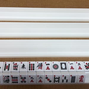 May include: A set of white Mahjong tile racks and tiles. The racks are rectangular with a lip to hold the tiles. The tiles are white with black and red symbols and numbers. The set includes four racks and a row of tiles.