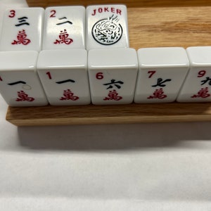 May include: A set of white mahjong tiles with red Chinese characters and numbers on them. The tiles are arranged in a row on a wooden surface. The tiles include numbers 1, 2, 3, 6, 7, and 9, as well as a Joker tile.